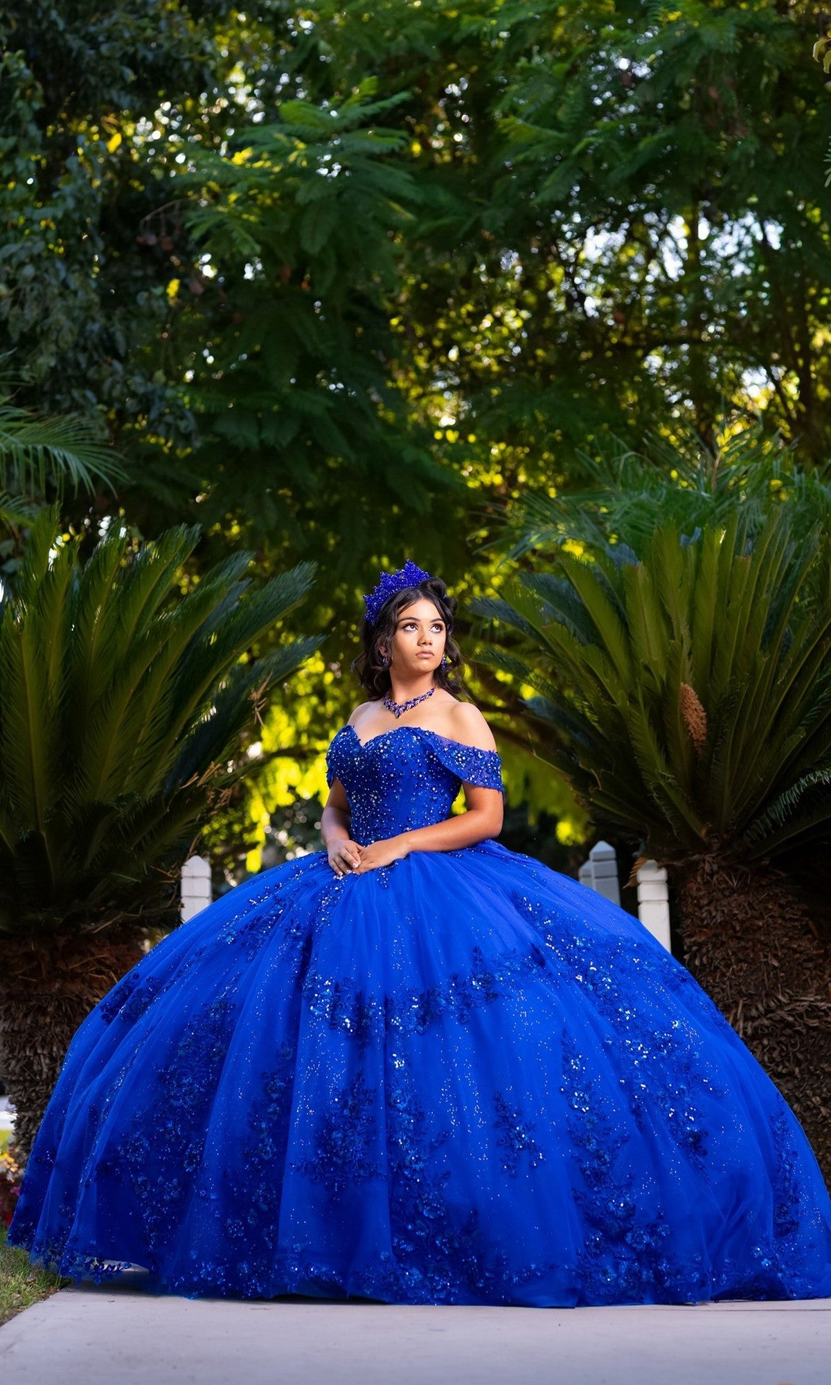 A woman stands outdoors among lush green plants, looking to the side in the stunning Quinceanera Dress CHF3122 By Chicas. The elegant off-the-shoulder design of this royal-blue gown highlights her graceful pose and creates a striking scene.